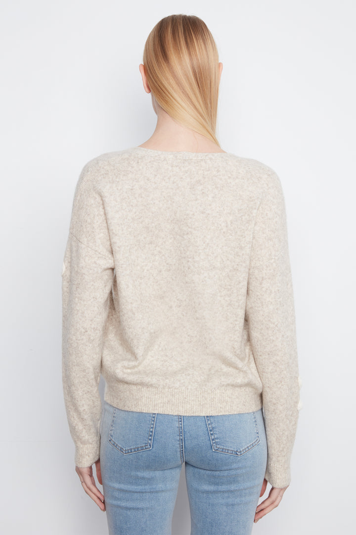 A woman shown from the back wearing a long sleeve beige sweater with cream embroidered flowers on the front. She is wearing blue jeans and standing in front of a light grey wall. 