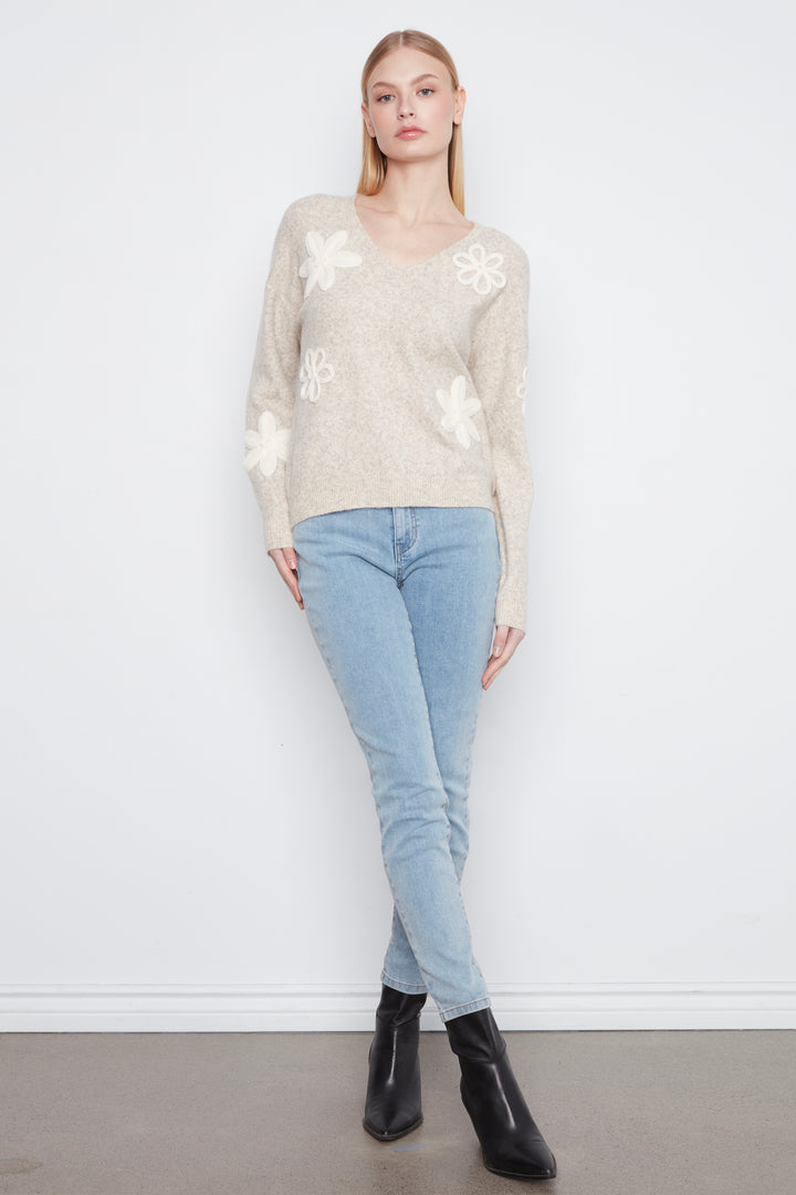 A woman wearing a long sleeve beige sweater with cream embroidered flowers on the front. She is wearing blue jeans and standing in front of a light grey wall. 