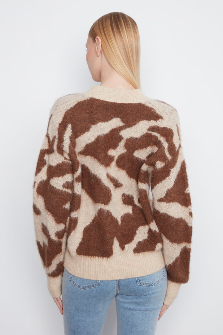 A woman shown from the back wearing a brown and beige camo print cardigan with front buttons. She is wearing blue jeans and standing in front of a light grey wall.