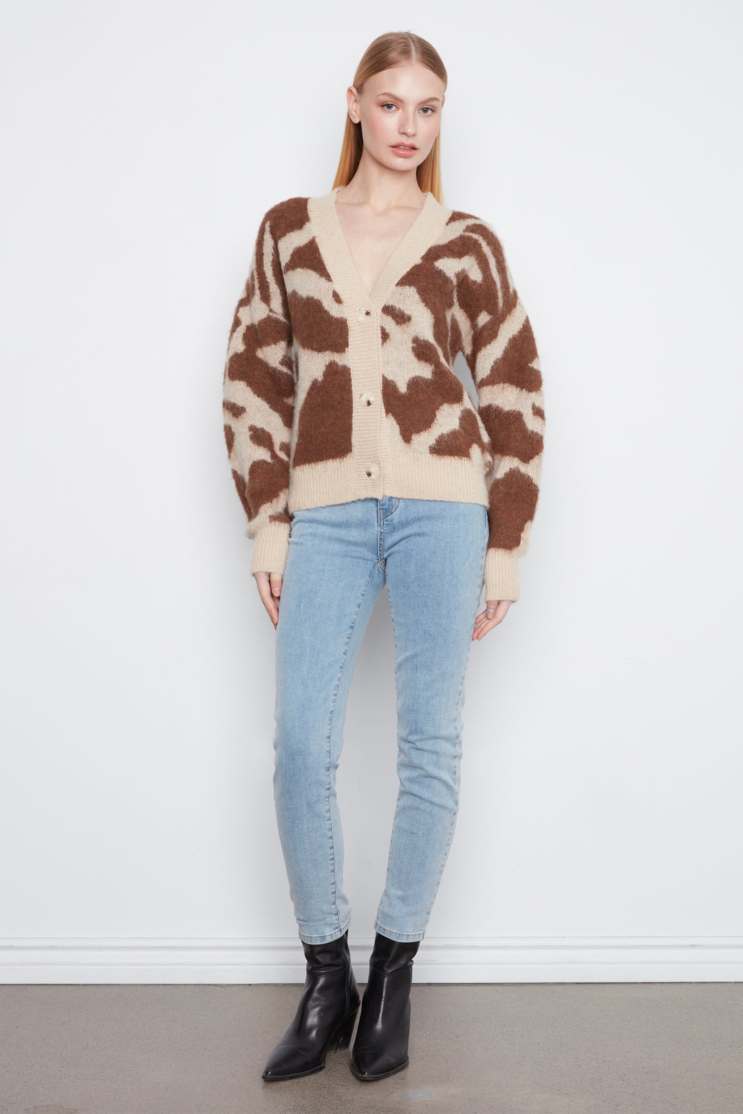 A woman wearing a brown and beige camo print cardigan with front buttons. She is wearing blue jeans and standing in front of a light grey wall.