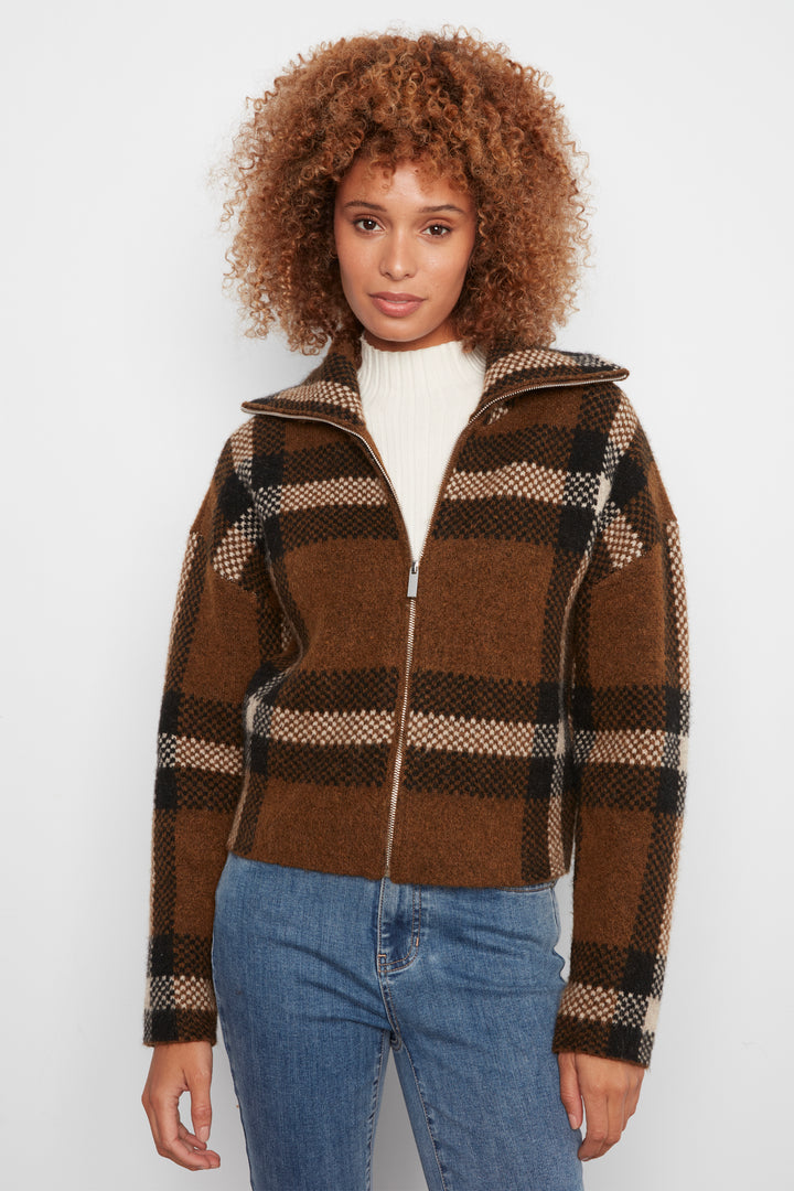 A woman wearing a brown plaid cropped cardigan and blue jeans standing in front of a white wall. 
