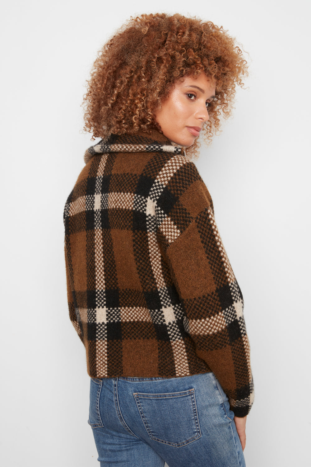 A woman shown from the back wearing a brown plaid cropped cardigan and blue jeans standing in front of a white wall. 