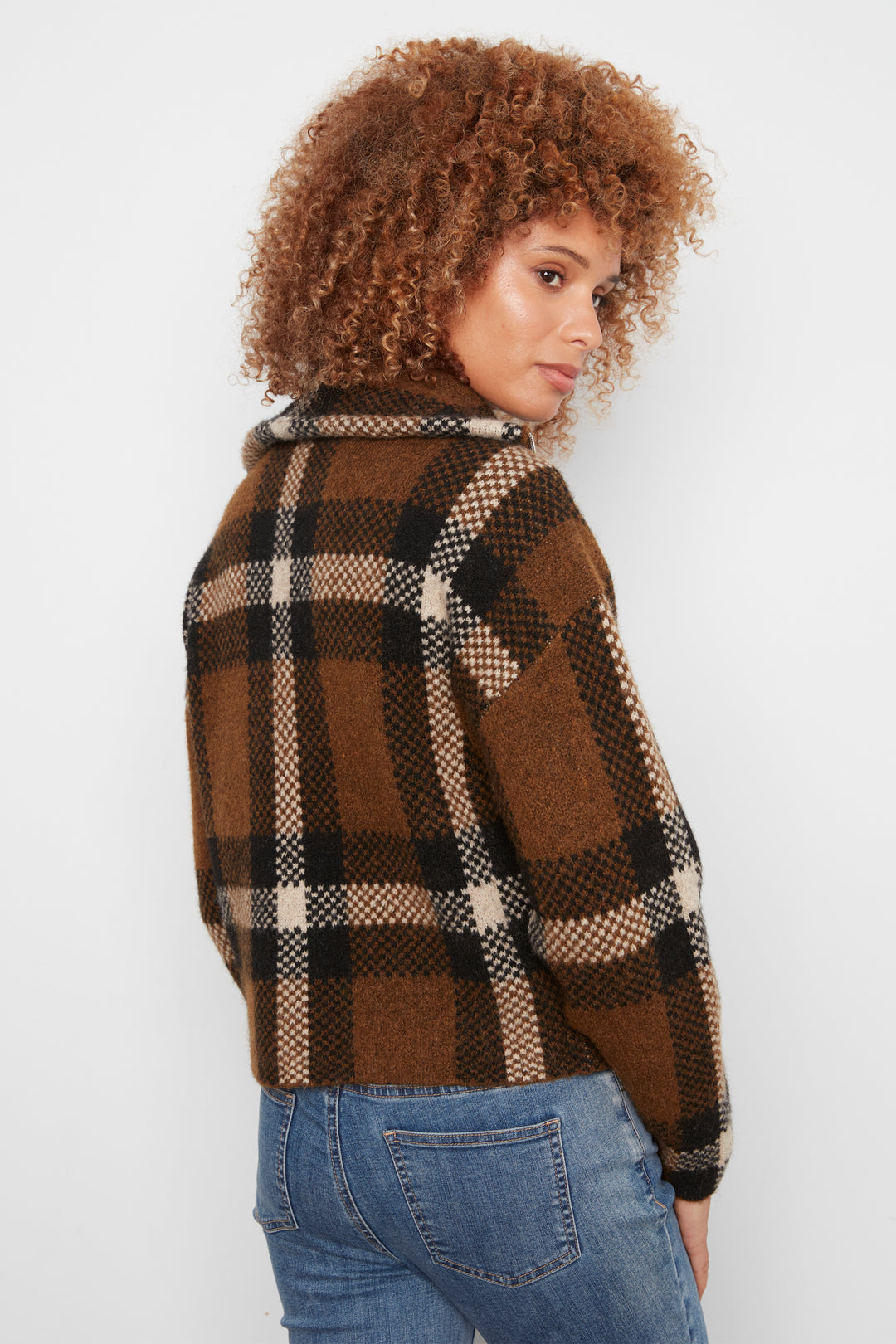 A woman shown from the back wearing a brown plaid cropped cardigan and blue jeans standing in front of a white wall. 