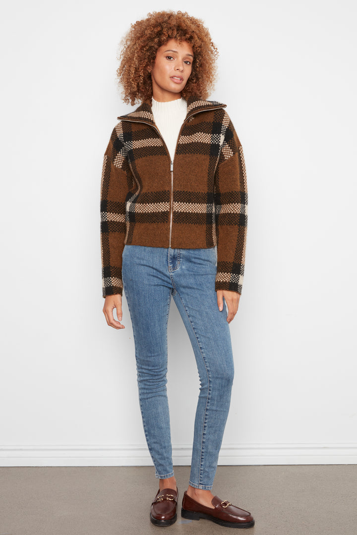 A woman wearing a brown plaid cropped cardigan and blue jeans standing in front of a white wall. 