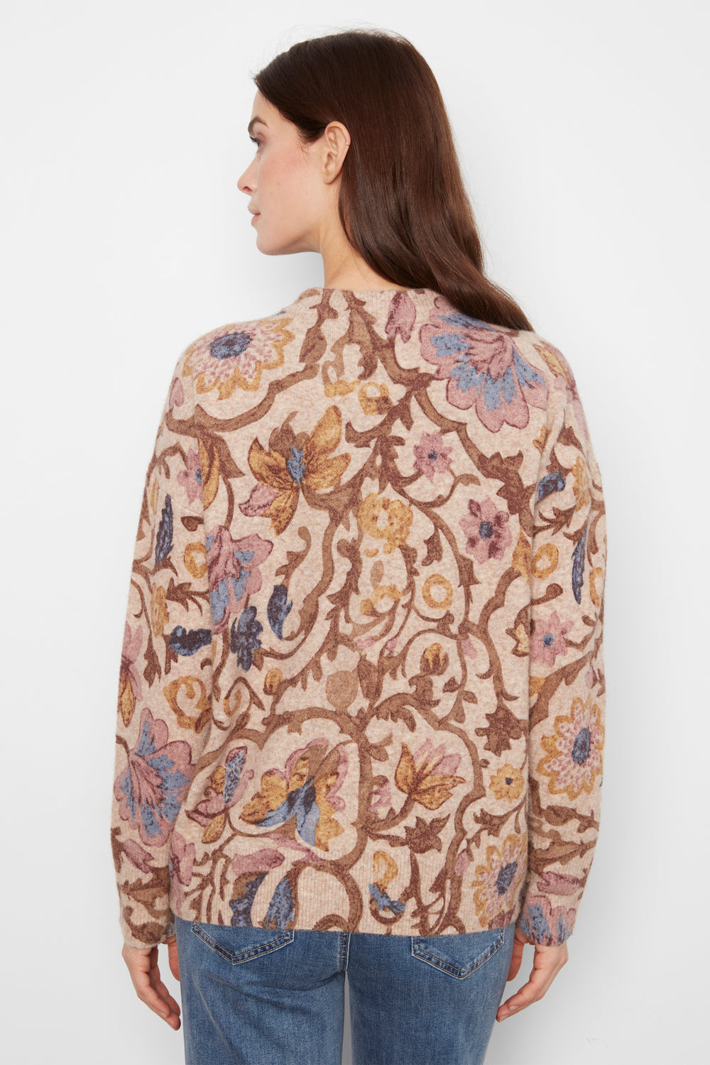 A woman shown from the back wearing a long sleeve baggy floral print sweater and jeans standing in front of a white wall. 