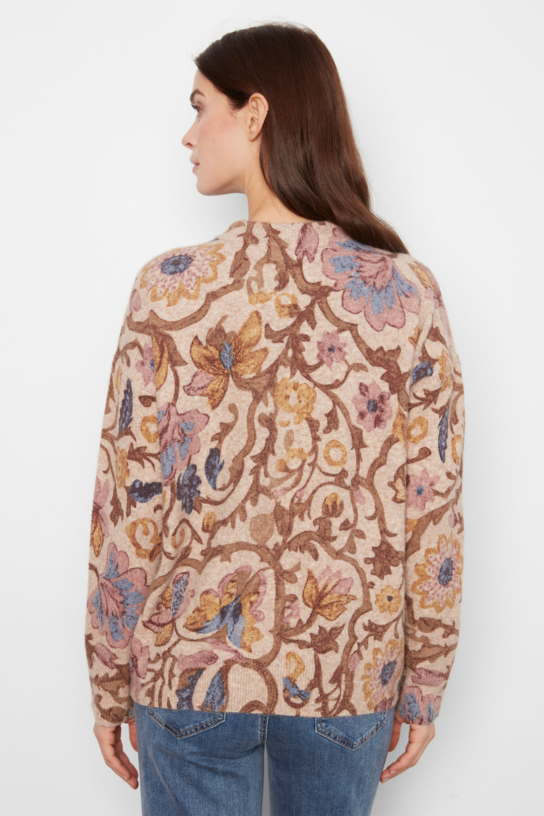 A woman shown from the back wearing a long sleeve baggy floral print sweater and jeans standing in front of a white wall. 