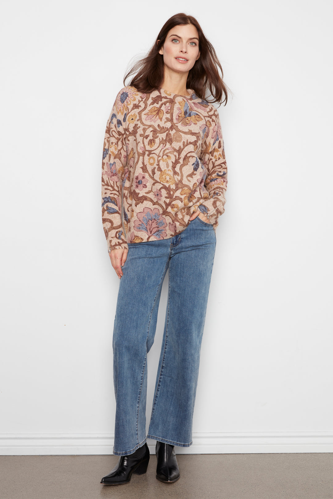 A woman wearing a long sleeve baggy floral print sweater and jeans standing in front of a white wall. 