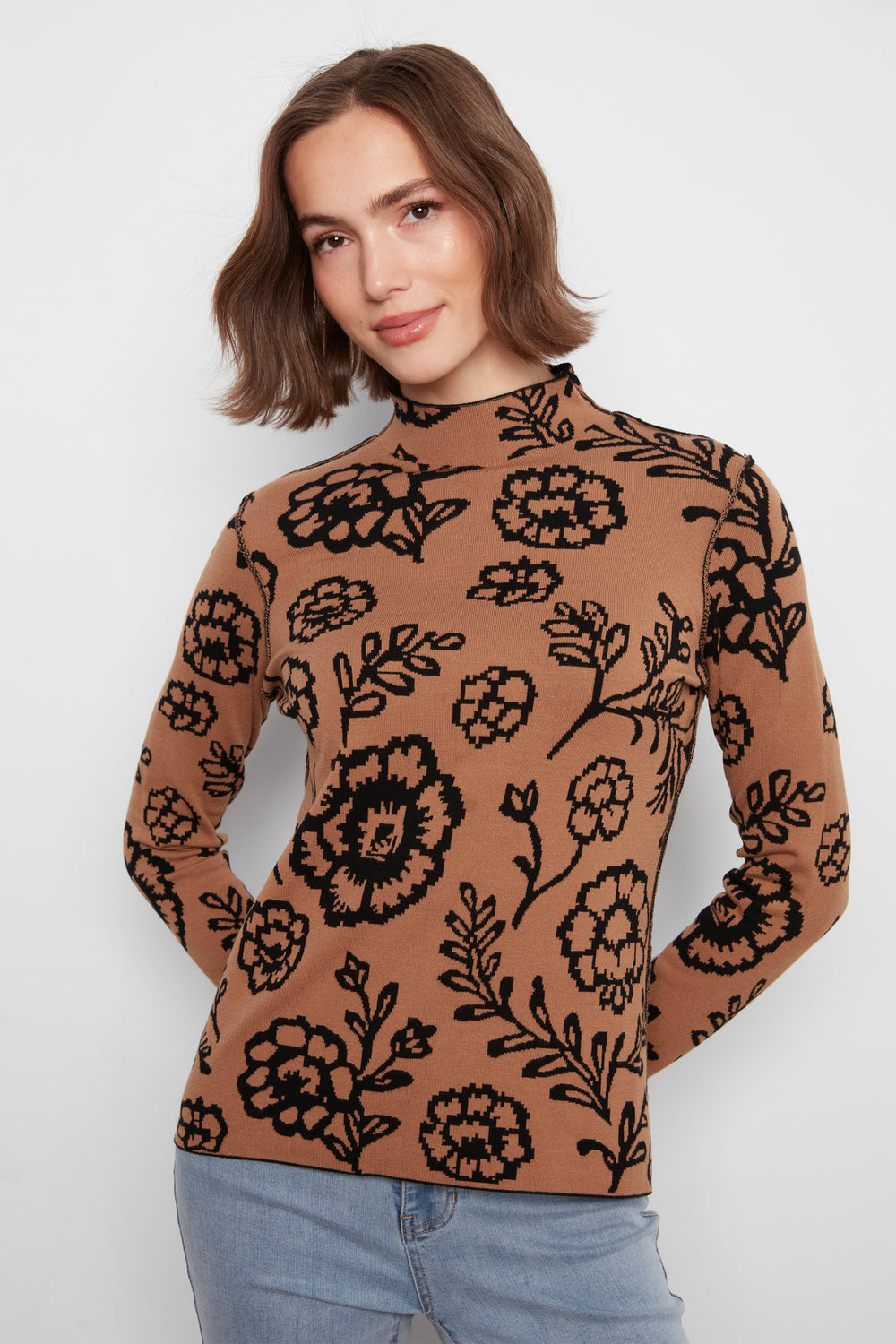 A woman wearing a mock neck long sleeve sweater with an all over floral print. She is wearing jeans and standing in front of a white wall. 
