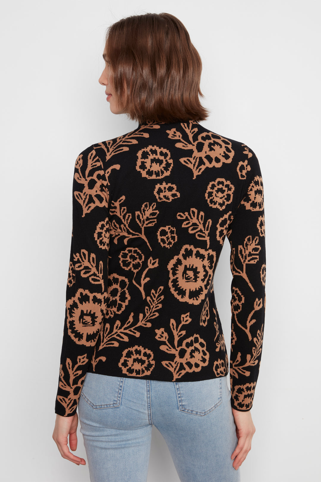 A woman shown from the back wearing a mock neck long sleeve sweater with an all over floral print. She is wearing jeans and standing in front of a white wall. 