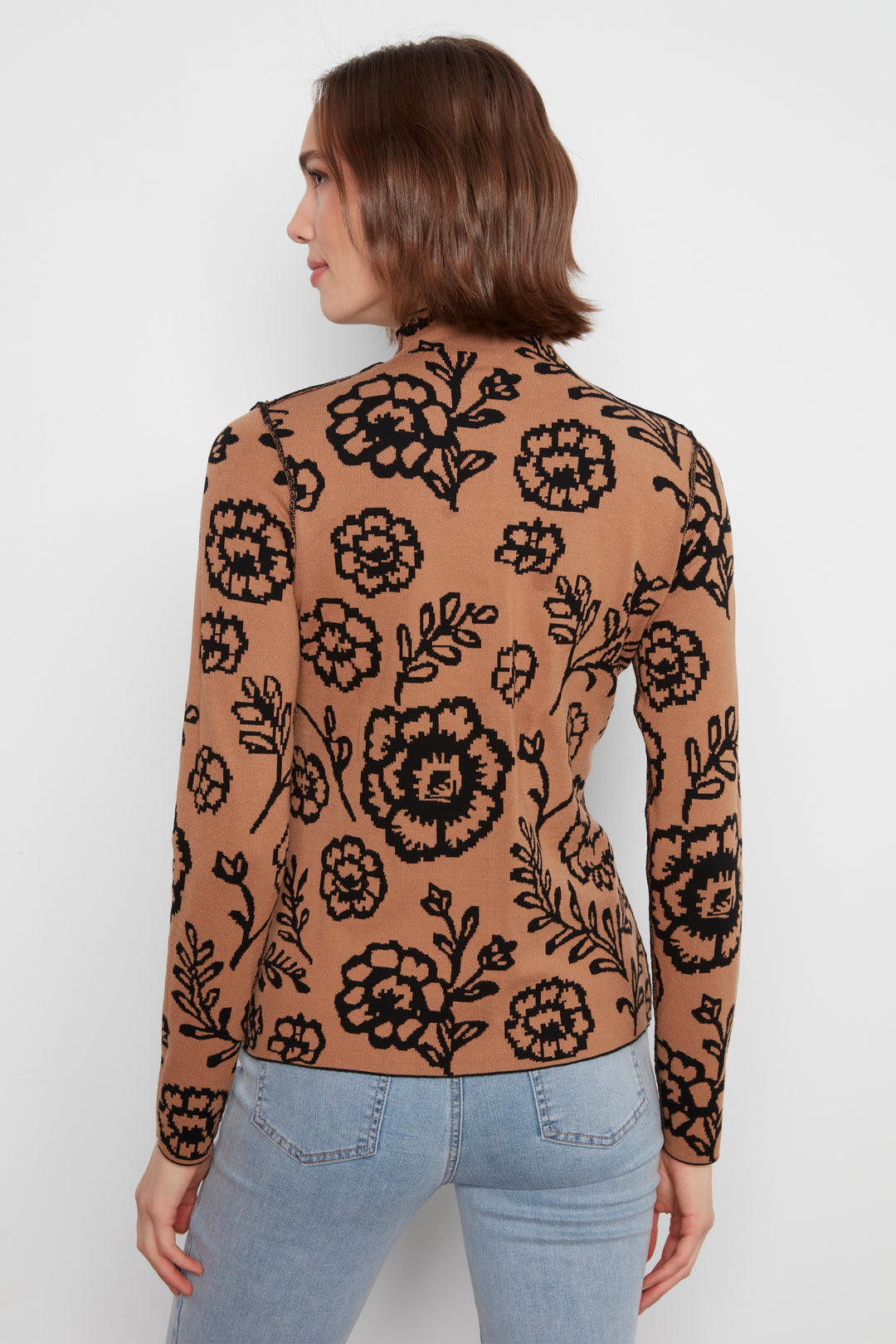 A woman shown from the back wearing a mock neck long sleeve sweater with an all over floral print. She is wearing jeans and standing in front of a white wall. 