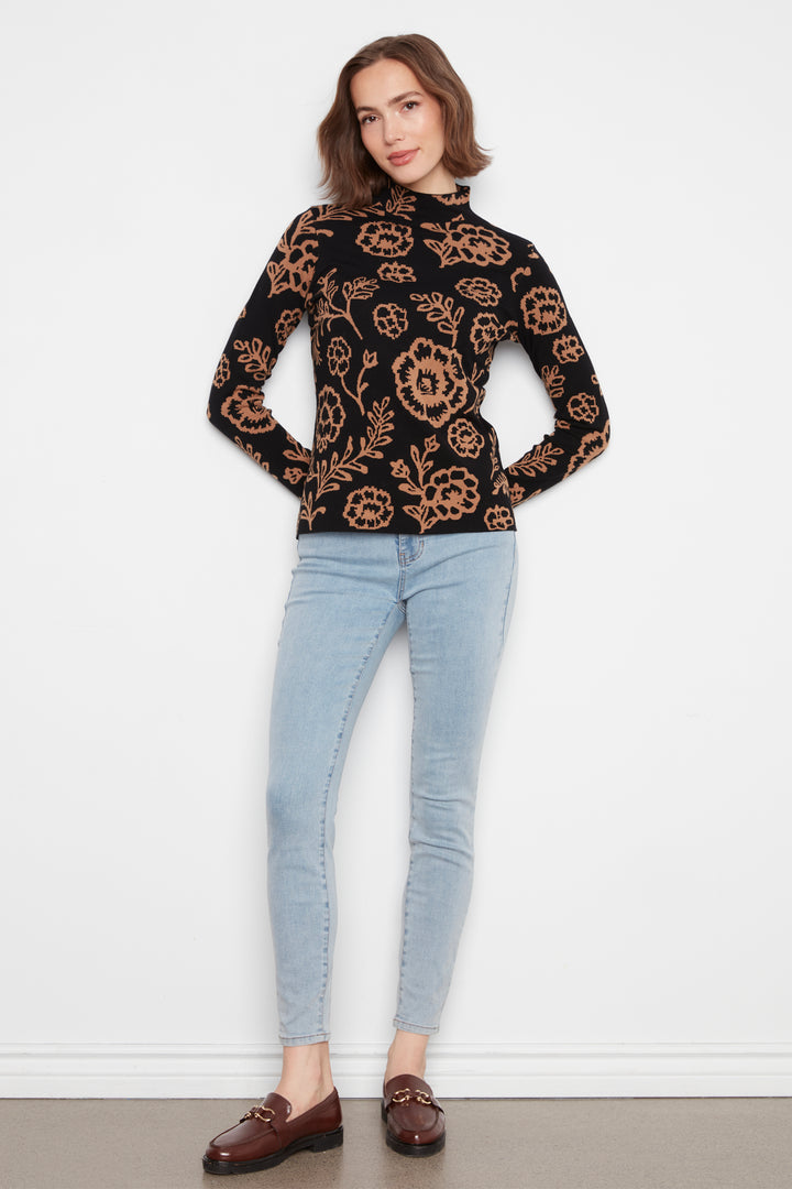 A woman wearing a mock neck long sleeve sweater with an all over floral print. She is wearing jeans and standing in front of a white wall. 