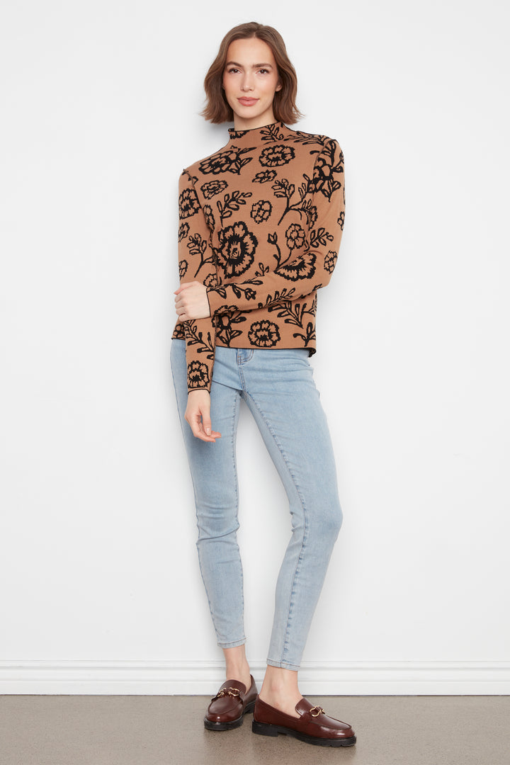 A woman wearing a mock neck long sleeve sweater with an all over floral print. She is wearing jeans and standing in front of a white wall. 