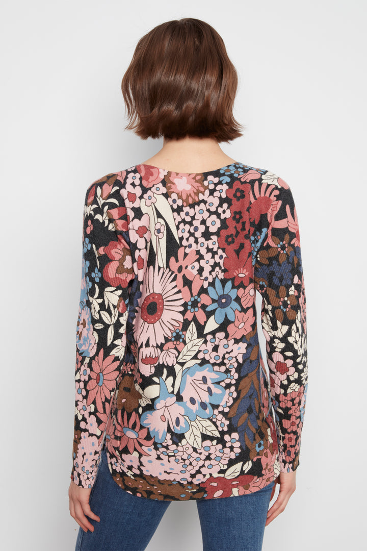 A woman shown from the back wearing a long sleeve flower print sweater top standing in front of a white wall. 