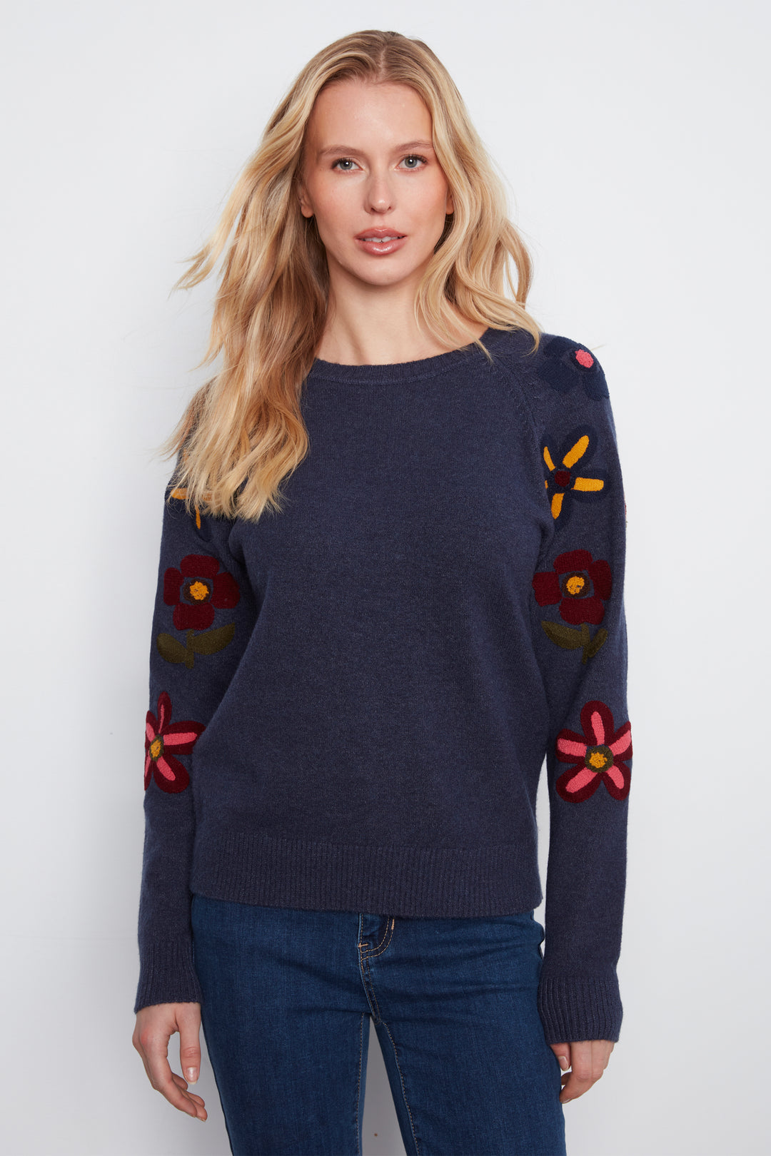 A woman wearing a navy long sleeve sweater with embroidered flowers on the sleeves standing in front of a white wall. 