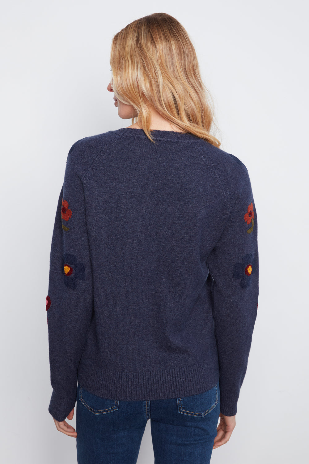 A woman shown from the back wearing a navy long sleeve sweater with embroidered flowers on the sleeves standing in front of a white wall. 