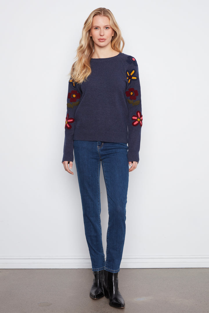 A woman wearing a navy long sleeve sweater with embroidered flowers on the sleeves standing in front of a white wall. 
