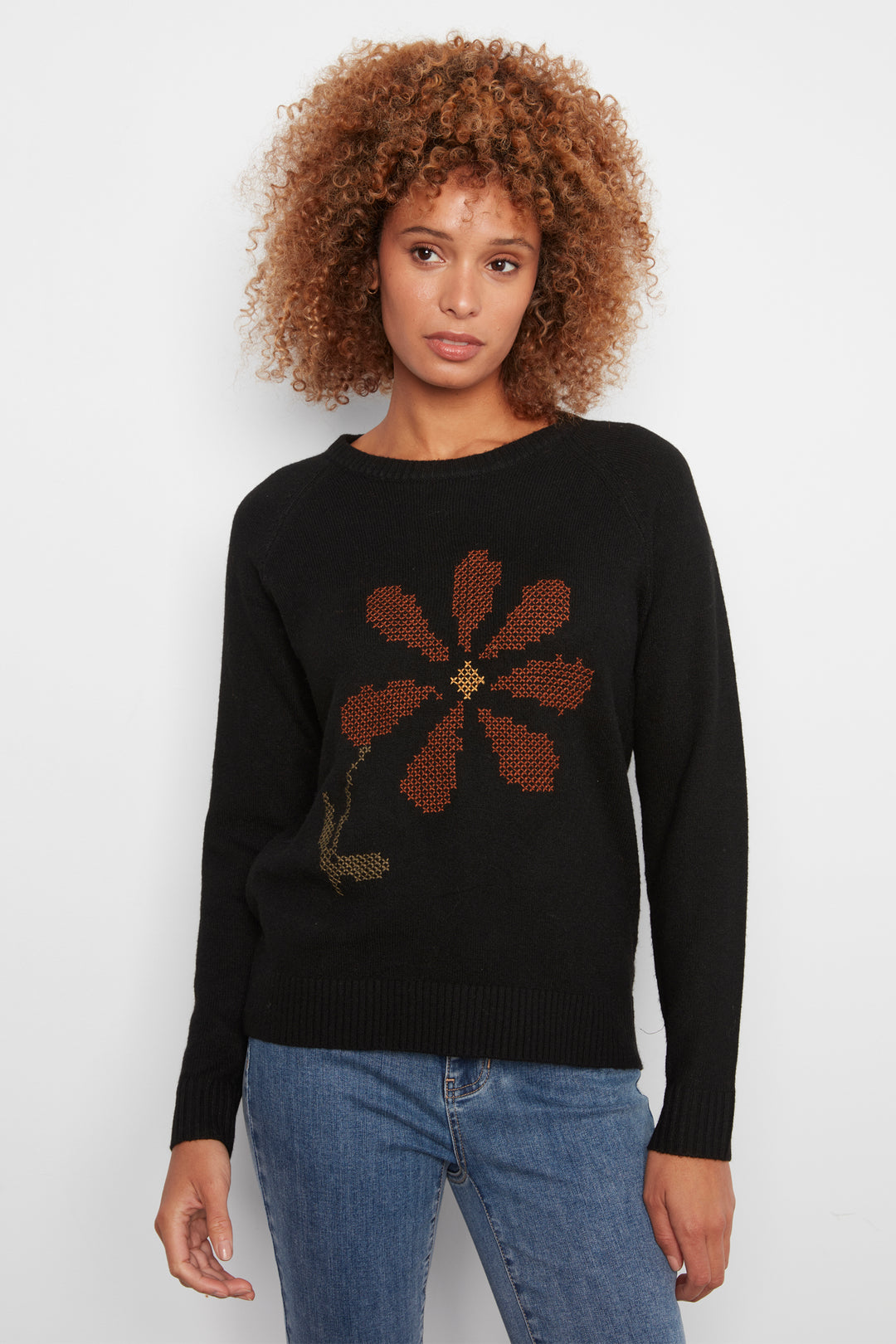 A woman wearing a black long sleeve sweater with a large flower on the front. She is wearing jeans and standing in front of a white wall. 