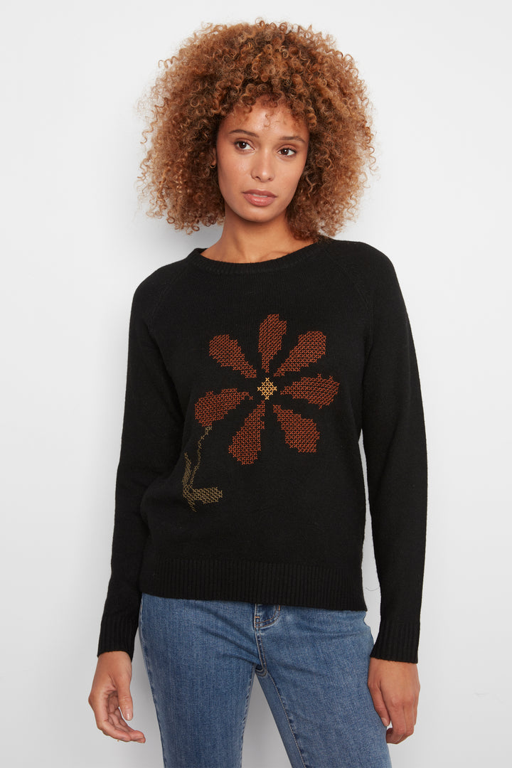 A woman wearing a black long sleeve sweater with a large flower on the front. She is wearing jeans and standing in front of a white wall. 