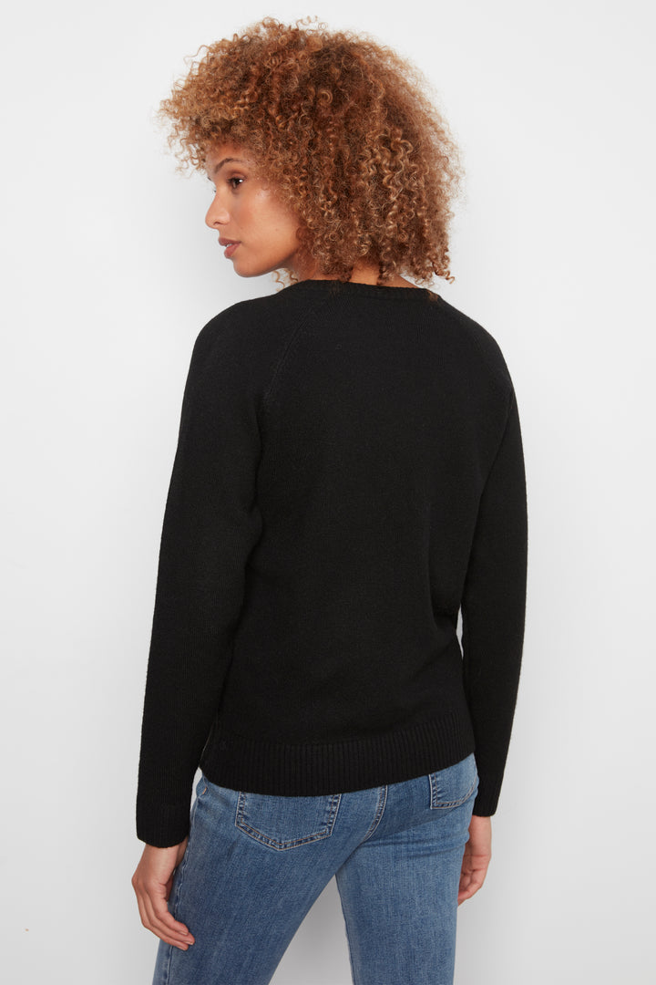 A woman shown from the back wearing a black long sleeve sweater with a large flower on the front. She is wearing jeans and standing in front of a white wall. 