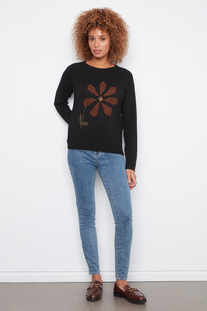 A woman wearing a black long sleeve sweater with a large flower on the front. She is wearing jeans and standing in front of a white wall. 