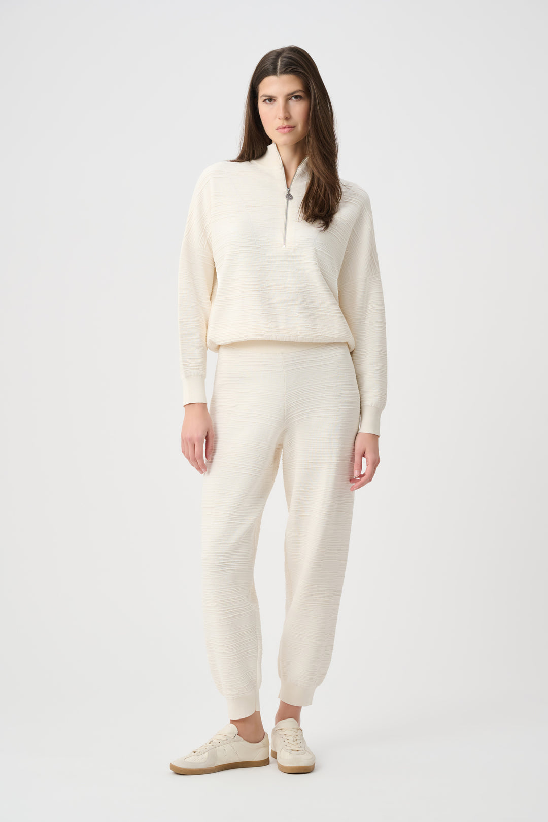 A woman wearing a cream half-zip textured sweatshirt with matching pants standing in front of a white background. 