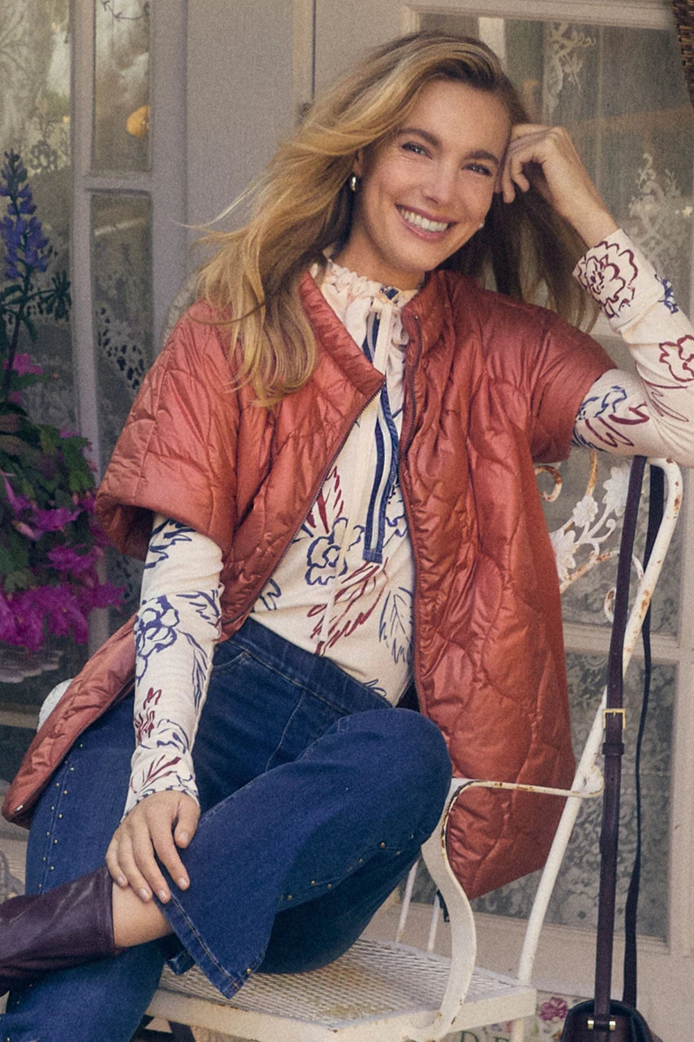 Woman wearing a red vest over a floral blouse and blue jeans, sitting on a chair.
