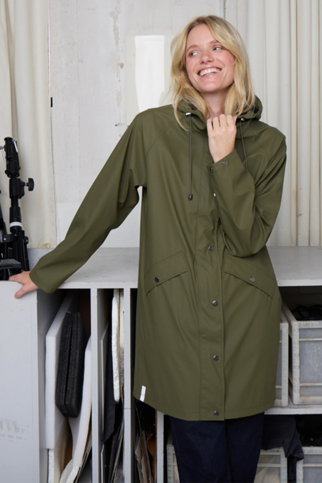 Woman wearing a green raincoat indoors with a white background