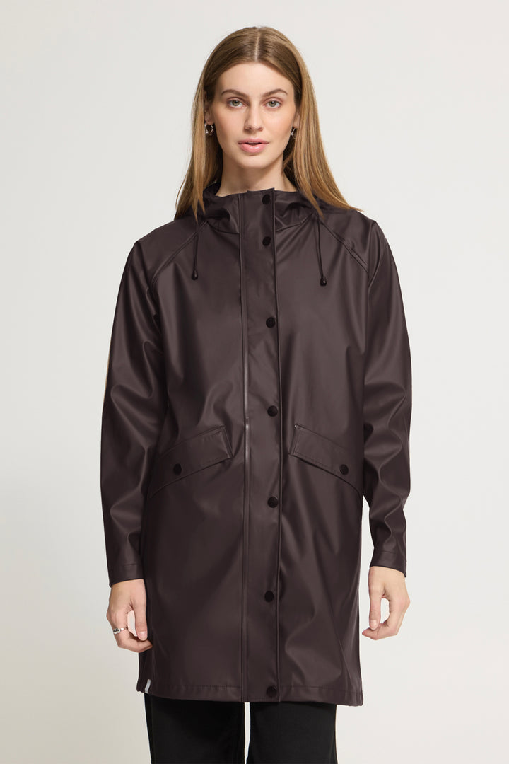 A woman modeling a dark brown hooded raincoat with a button-front closure and flap pockets, photographed against a plain light gray studio background.