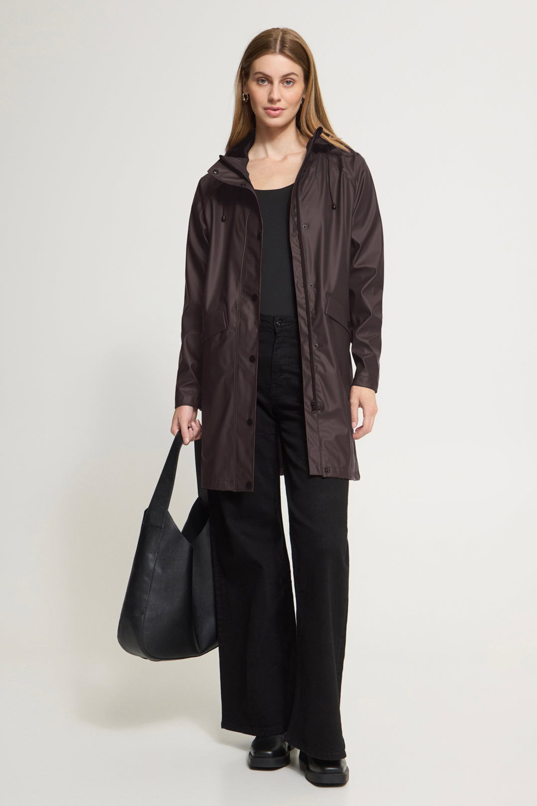 A woman stands against a light gray studio background wearing a dark brown hooded raincoat over a black outfit, holding a large black tote bag.