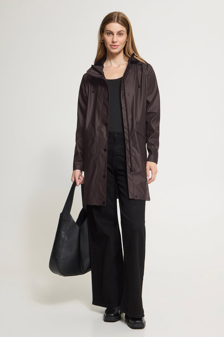 A woman stands against a light gray studio background wearing a dark brown hooded raincoat over a black outfit, holding a large black tote bag.