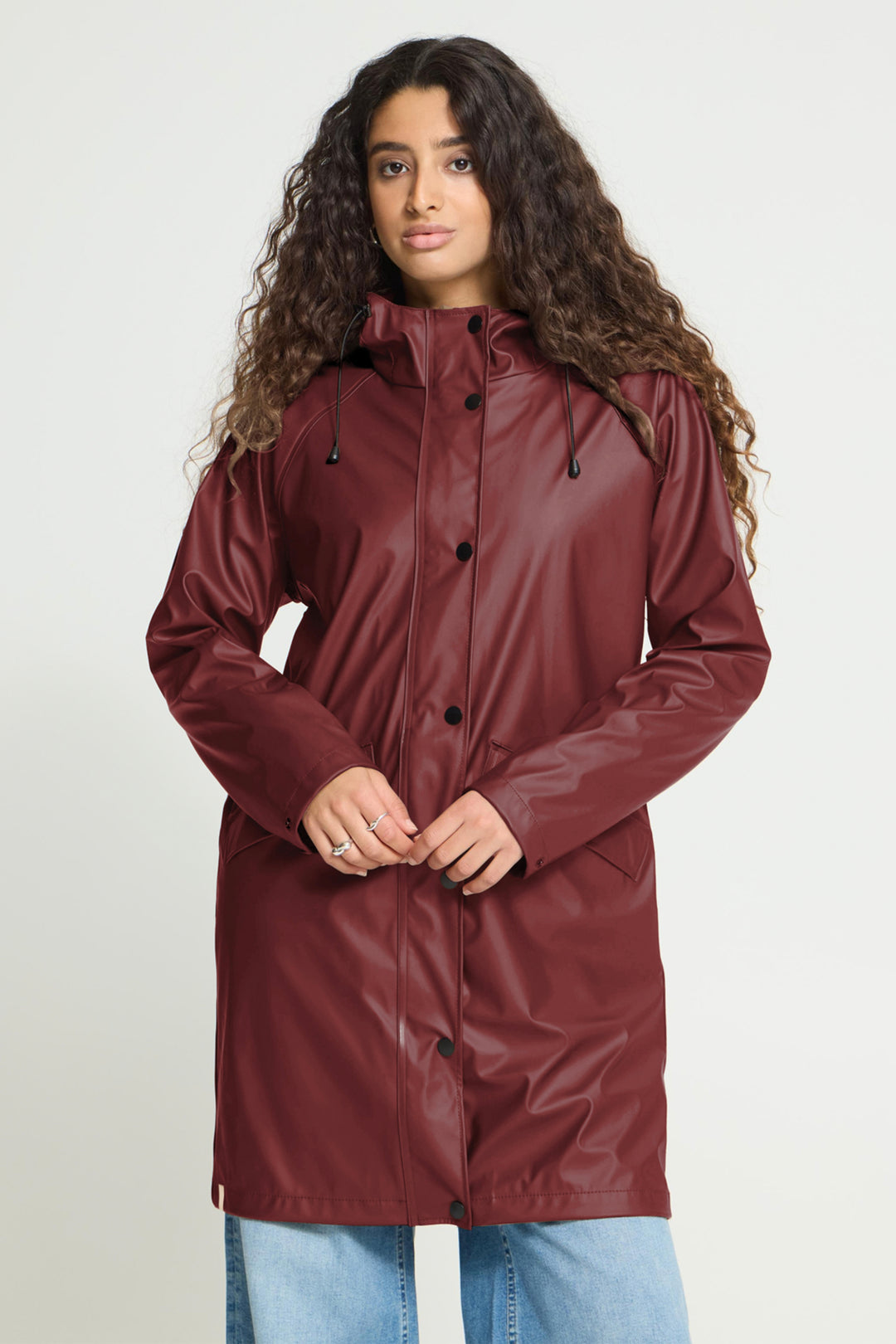 A woman with long curly brown hair wearing a glossy, long burgundy raincoat and light blue jeans, standing against a light gray studio background.