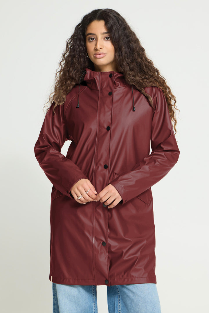 A woman with long curly brown hair wearing a glossy, long burgundy raincoat and light blue jeans, standing against a light gray studio background.