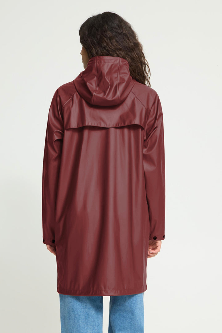 A woman viewed from the back wearing a hooded raincoat in a deep burgundy color, paired with blue jeans, against a plain light gray studio background.