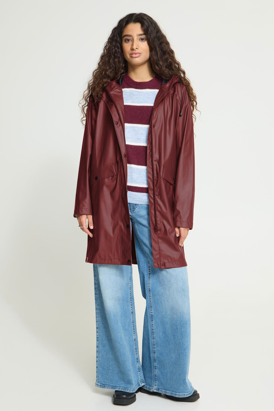 A model wearing a glossy burgundy hooded raincoat over a maroon and white striped sweater, paired with wide-leg light blue jeans and black shoes, against a plain light gray studio background.
