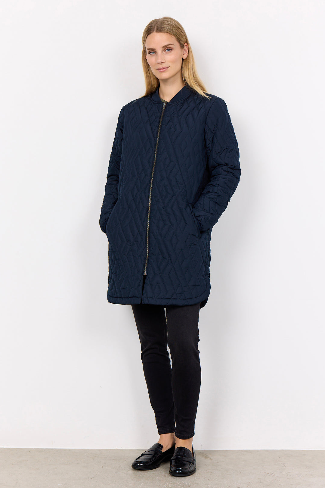 A woman modeling a dark navy blue quilted jacket with a front zipper and pockets, paired with black pants and black loafers, standing against a plain white wall.