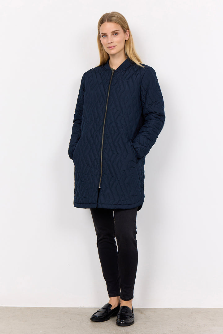 A woman modeling a dark navy blue quilted jacket with a front zipper and pockets, paired with black pants and black loafers, standing against a plain white wall.