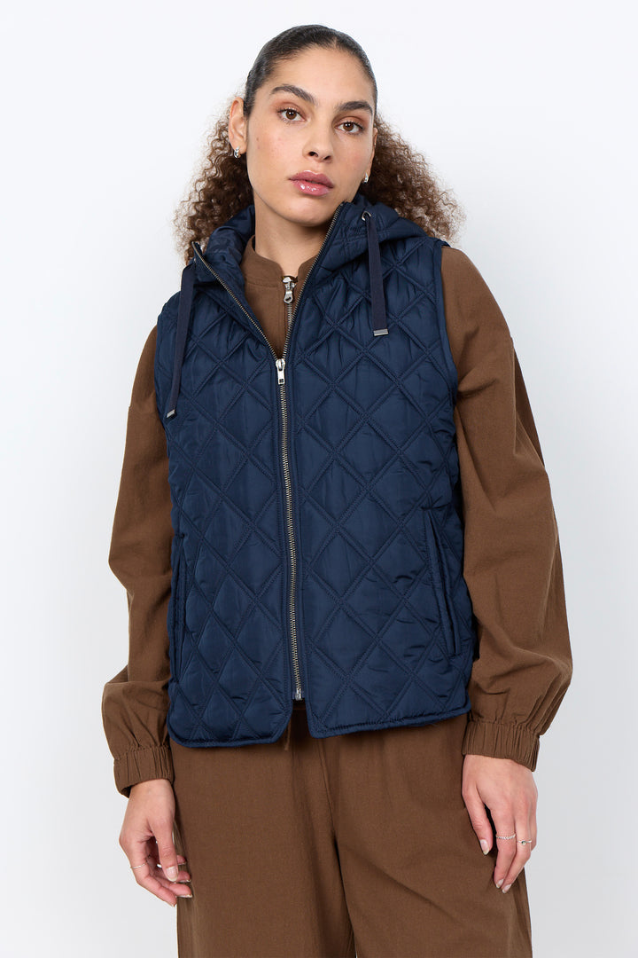 A model wearing a navy blue diamond-quilted zip-up puffer vest over a brown jacket and matching brown pants, photographed against a light gray studio background.