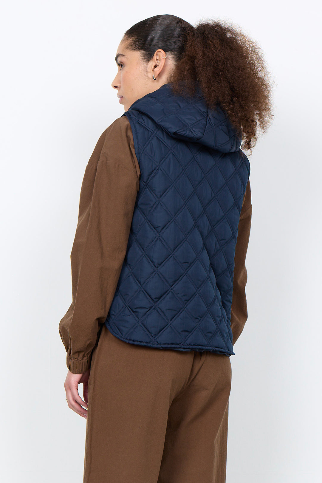 A woman shown from the back wearing a navy blue diamond-quilted hooded puffer vest over a brown jacket and brown pants, photographed against a plain light gray studio background.