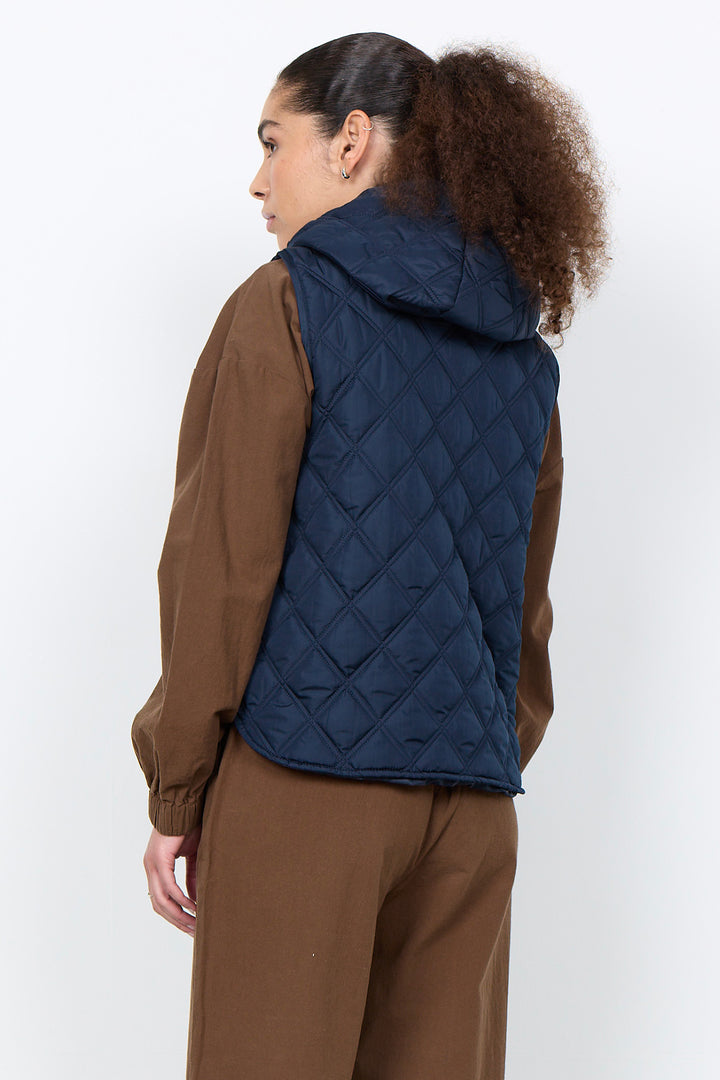 A woman shown from the back wearing a navy blue diamond-quilted hooded puffer vest over a brown jacket and brown pants, photographed against a plain light gray studio background.