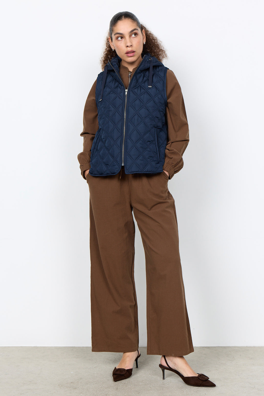 A woman stands against a plain light gray studio wall wearing a navy quilted zip-front puffer vest over a brown long-sleeve top and matching brown wide-leg pants, with dark pointed heels.