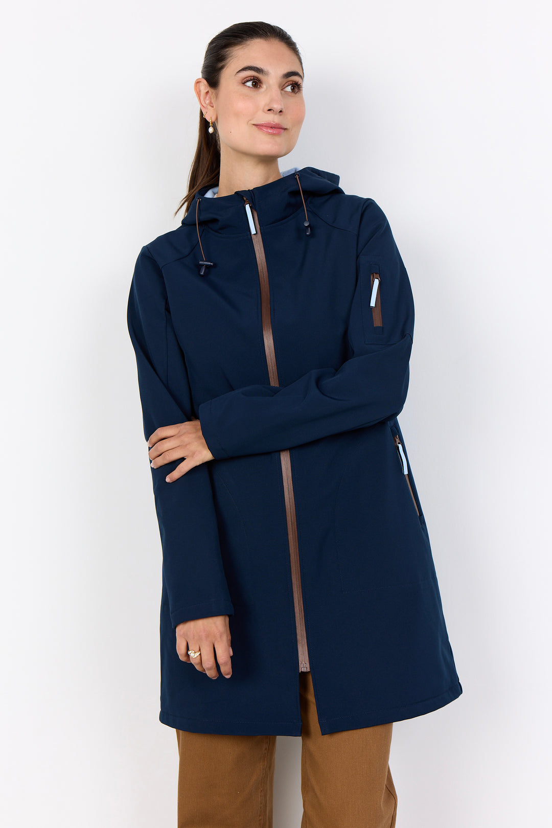 A woman wearing a navy blue hooded jacket with brown trim details, paired with brown pants, standing against a plain white studio background.