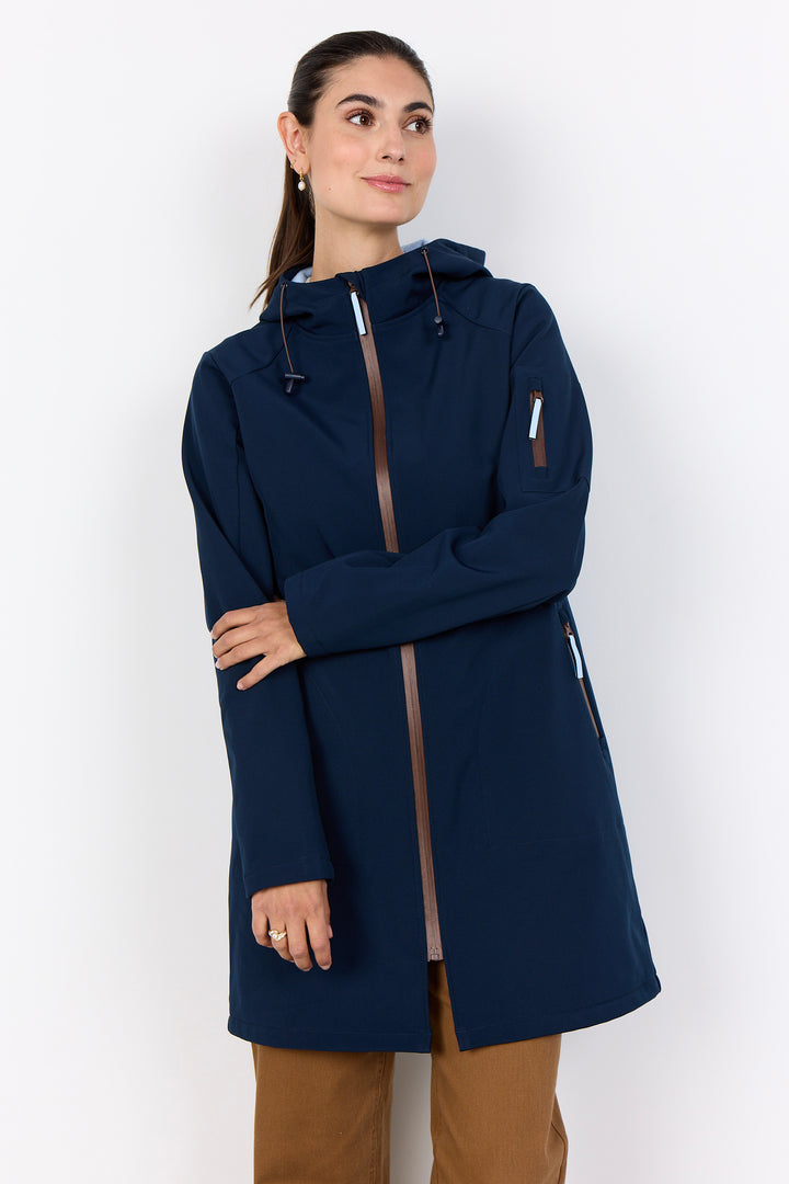 A woman wearing a navy blue hooded jacket with brown trim details, paired with brown pants, standing against a plain white studio background.