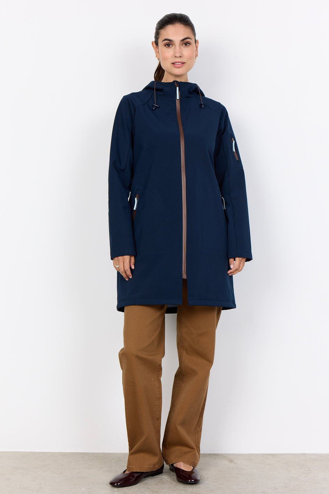 A woman modeling a navy blue hooded raincoat with a brown zipper, paired with brown trousers and dark brown shoes, standing against a plain white studio background.