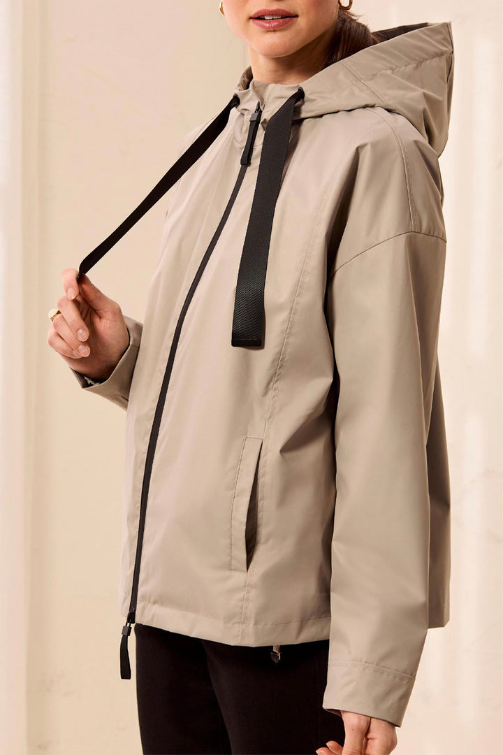 A woman wearing a light beige hooded jacket with black zippers and drawstrings, shown from the mouth down against a soft cream background.