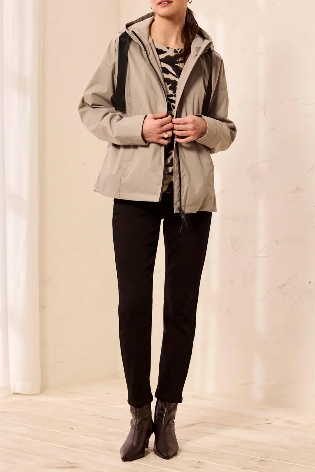 A woman wearing a light beige hooded jacket over a black and white patterned top, black pants, and dark brown ankle boots, standing in a brightly lit indoor setting with a cream wall and light wood floor.