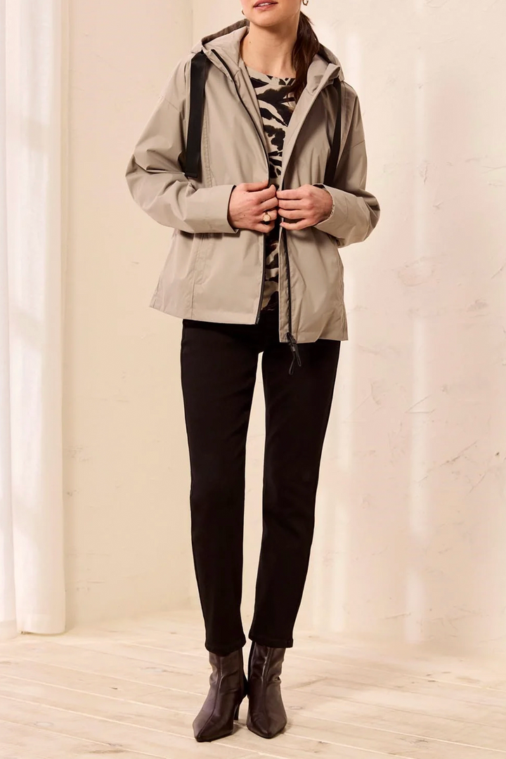 A woman wearing a light beige hooded jacket over a black and white patterned top, black pants, and dark brown ankle boots, standing in a brightly lit indoor setting with a cream wall and light wood floor.