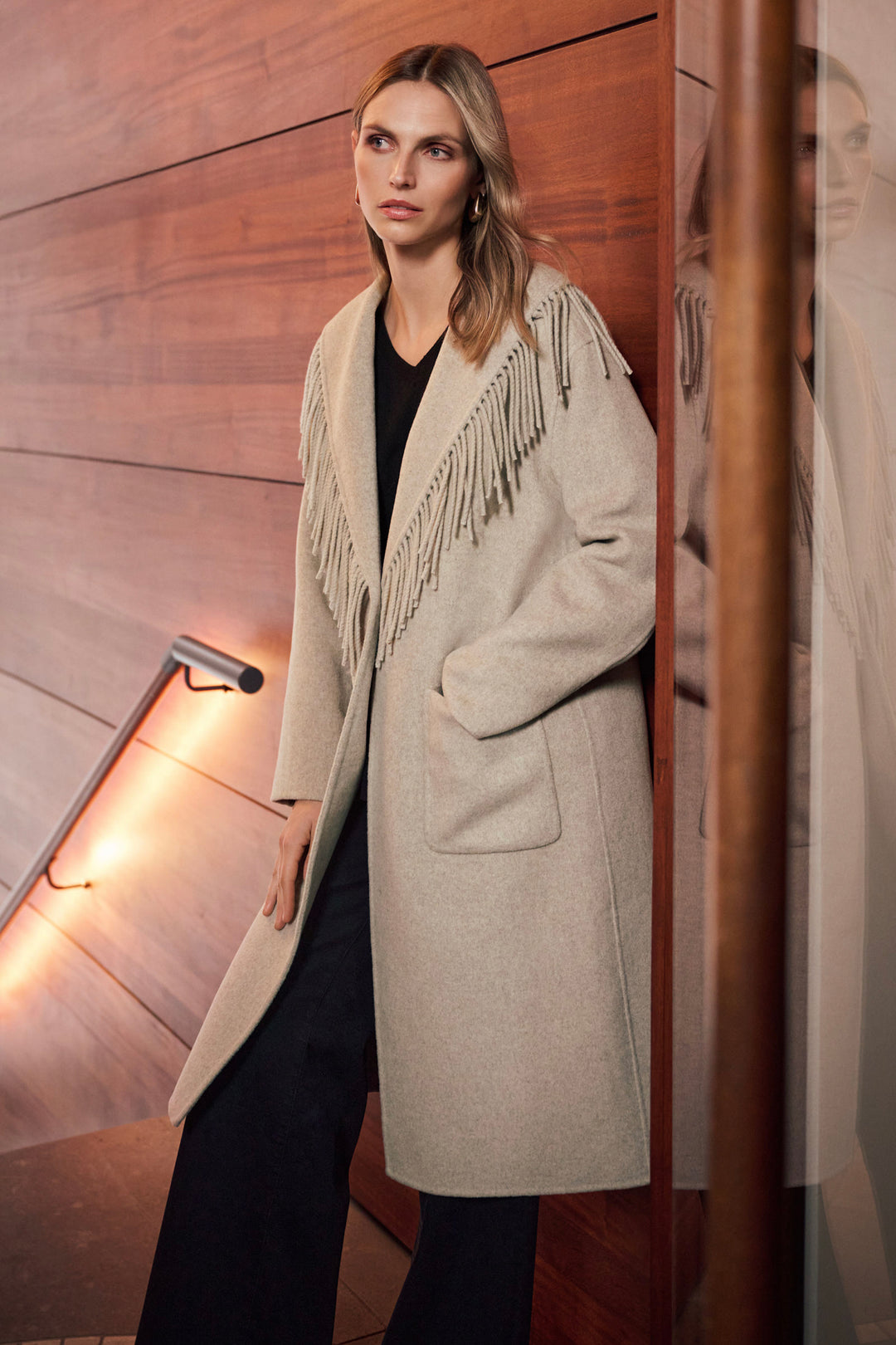 A woman wearing a long, light beige fringed coat over a black outfit, standing in front of a warm wood-paneled wall with a lit railing.