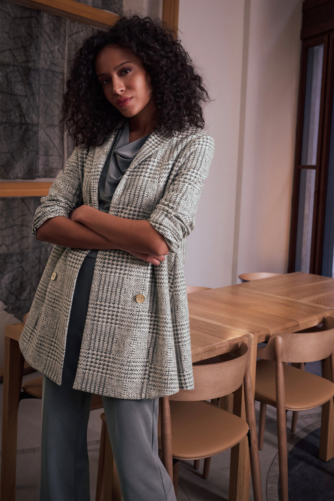 A woman with curly dark hair stands indoors with her arms crossed, wearing a light beige and gray houndstooth blazer over a gray top and gray pants. The setting includes a wooden dining table and chairs in a softly lit interior.