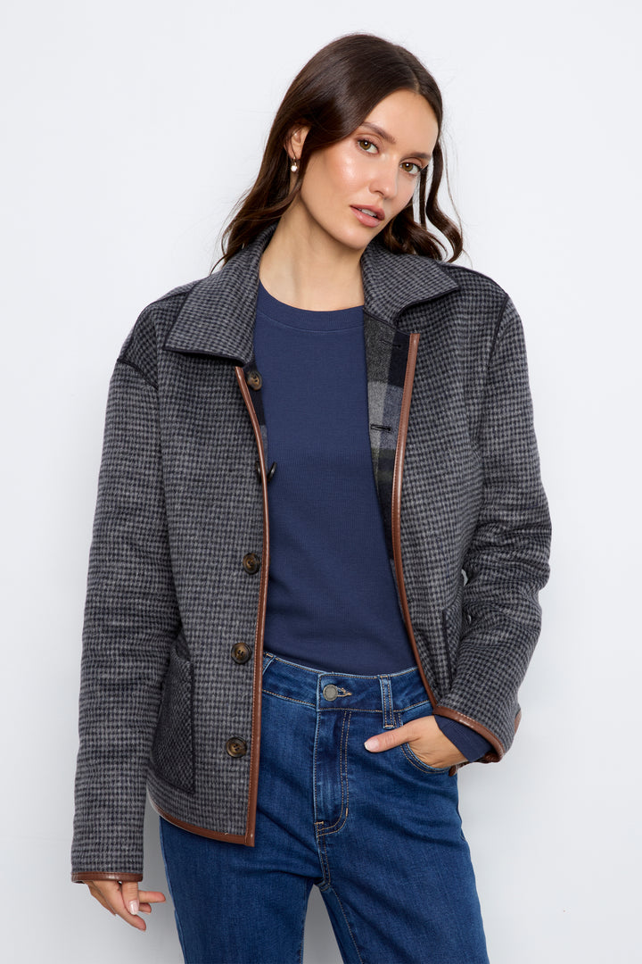 A woman modeling a dark gray and black houndstooth jacket with brown leather trim, worn over a navy blue top and blue jeans, against a light gray studio background.