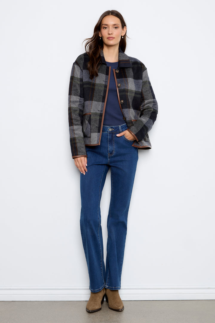 A woman modeling a dark gray and navy plaid textured jacket with brown buttons and brown trim, paired with blue jeans and brown ankle boots, standing against a white wall.